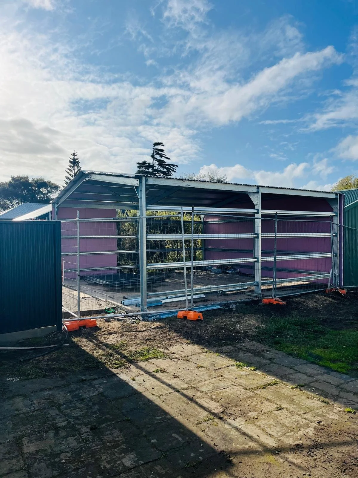 Construction site of a small purple structure with metal framing, surrounded by a temporary fence, under a partly cloudy sky.