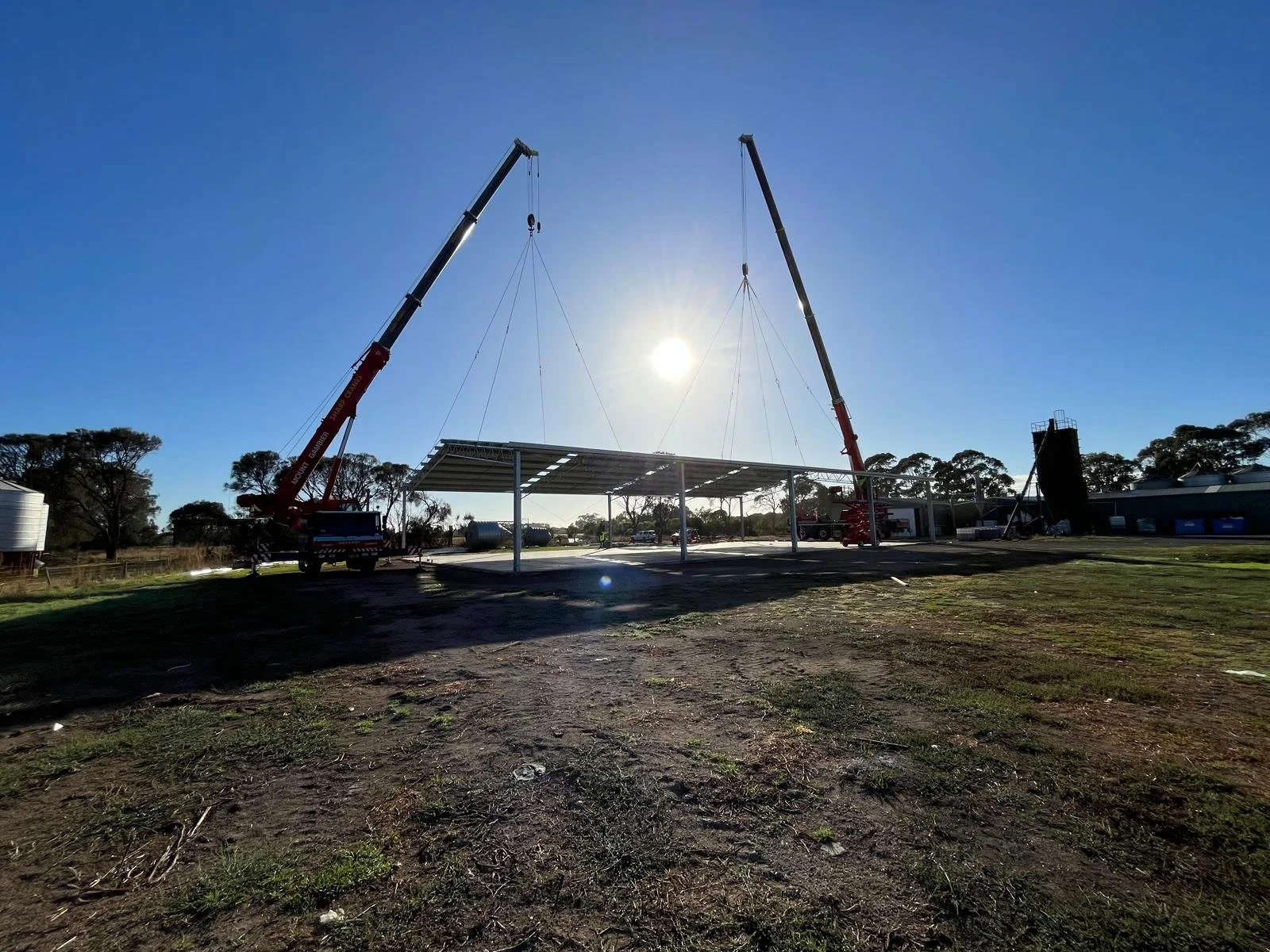Construction site with two cranes lifting a metal roof structure, set against a bright blue sky with the sun shining.