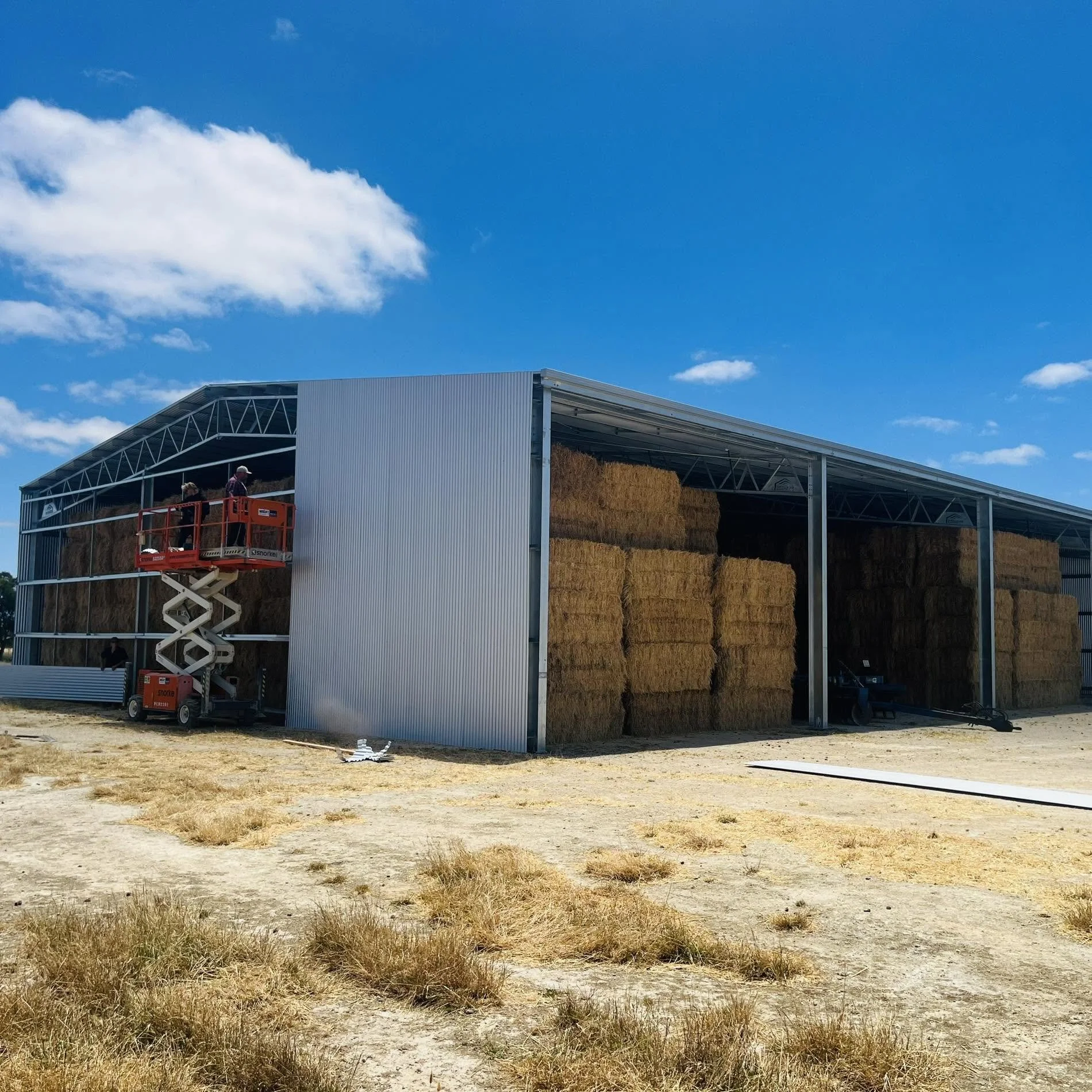A large open-sided metal structure holding hay bales, with people on a scissor lift working on the structure, under a partly cloudy blue sky with dry grass in the foreground.
