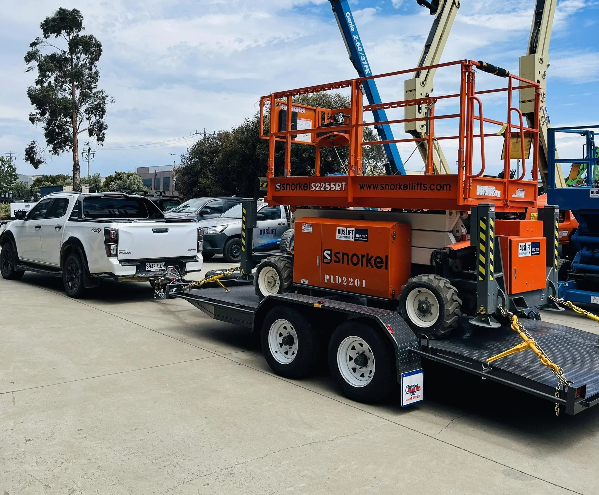 A construction lift parked on a parking lot with vehicles, trees, and a building in the background under a partly cloudy sky.