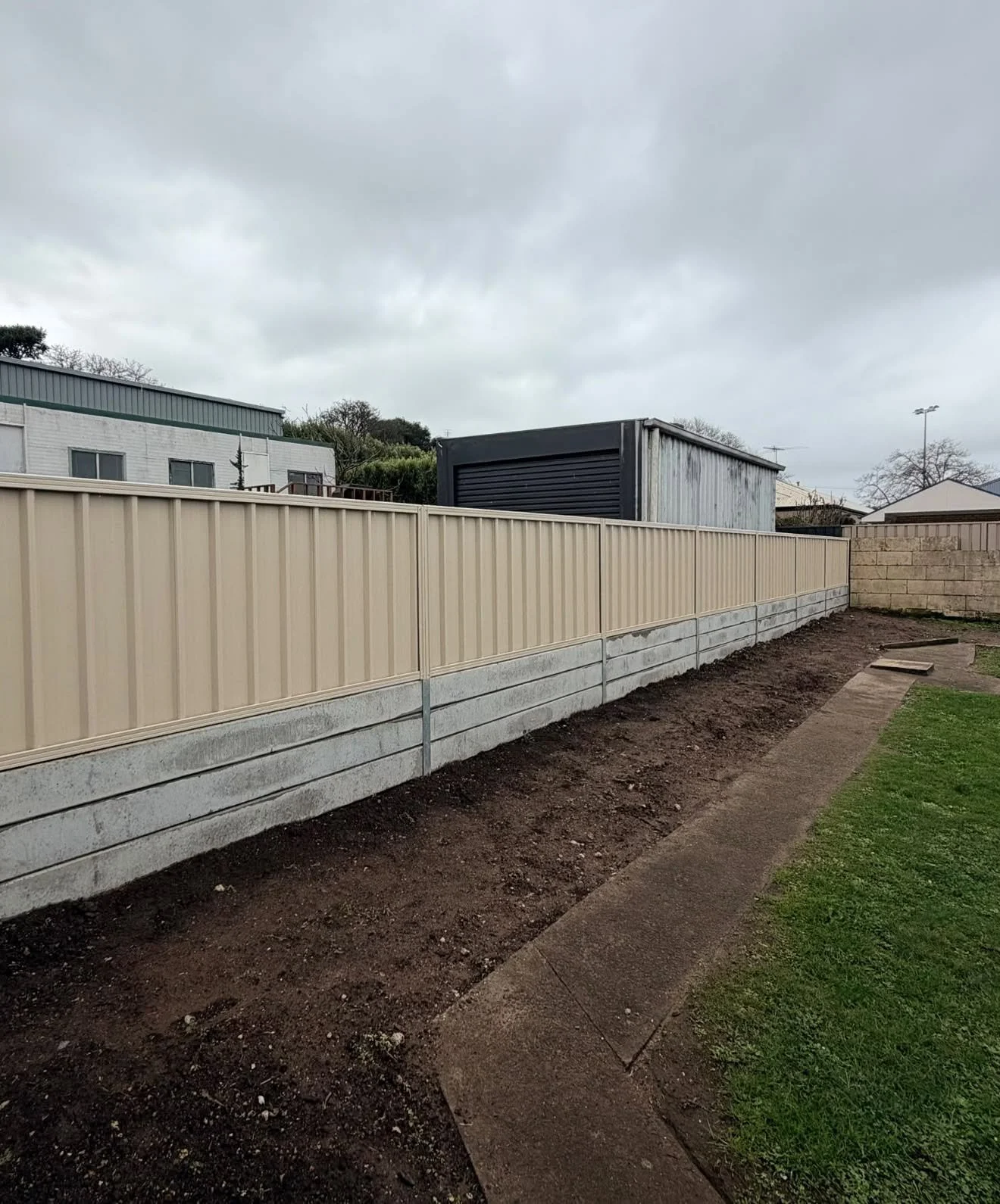 Backyard with a new cream-colored fence, a concrete base, and a narrow dirt pathway. A small grassy area is on the right. There are cloudy skies overhead.