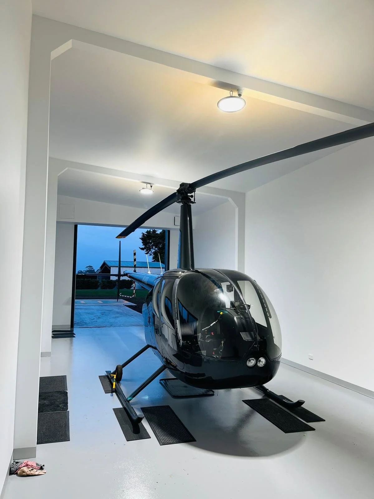 Black helicopter inside a modern, white-walled room with large window showing outdoor scenery, including trees and a building, during early evening or dawn.