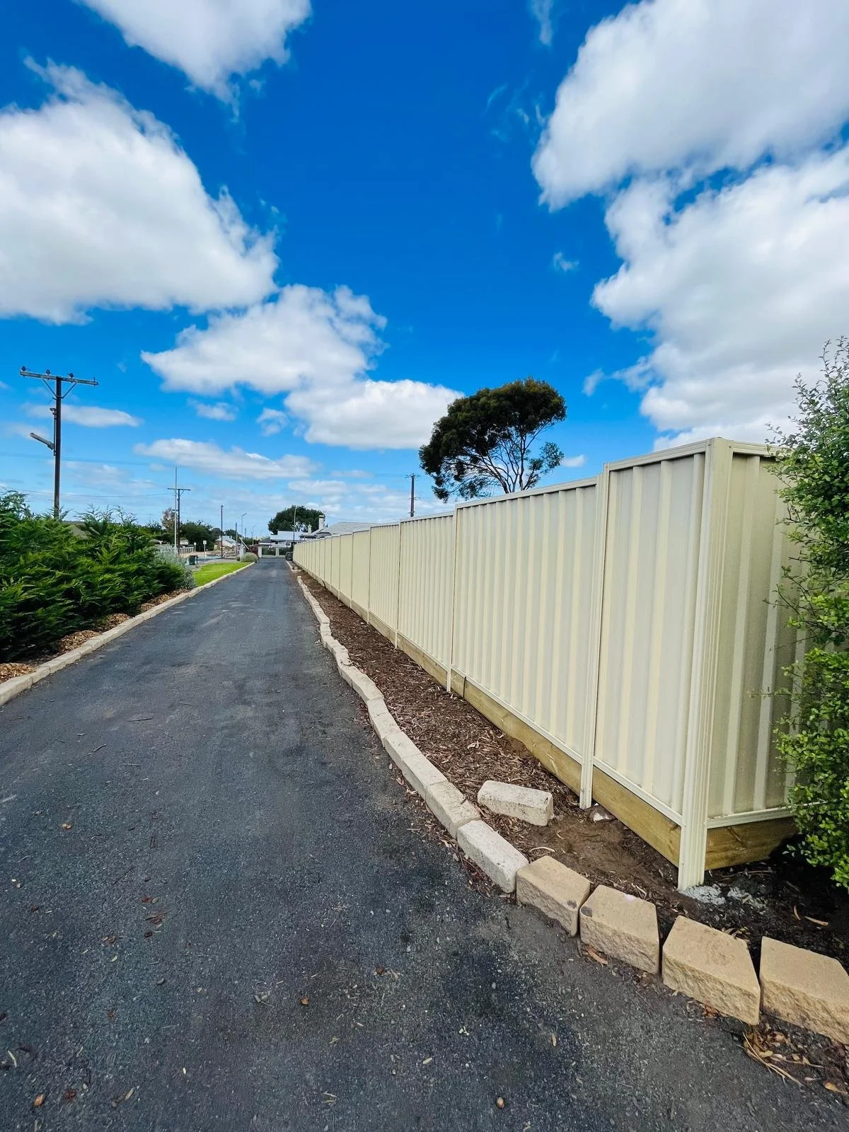A quiet street with a dirt path, green bushes on the left, a white fence on the right, a tall tree in the distance, and a blue sky with white clouds.
