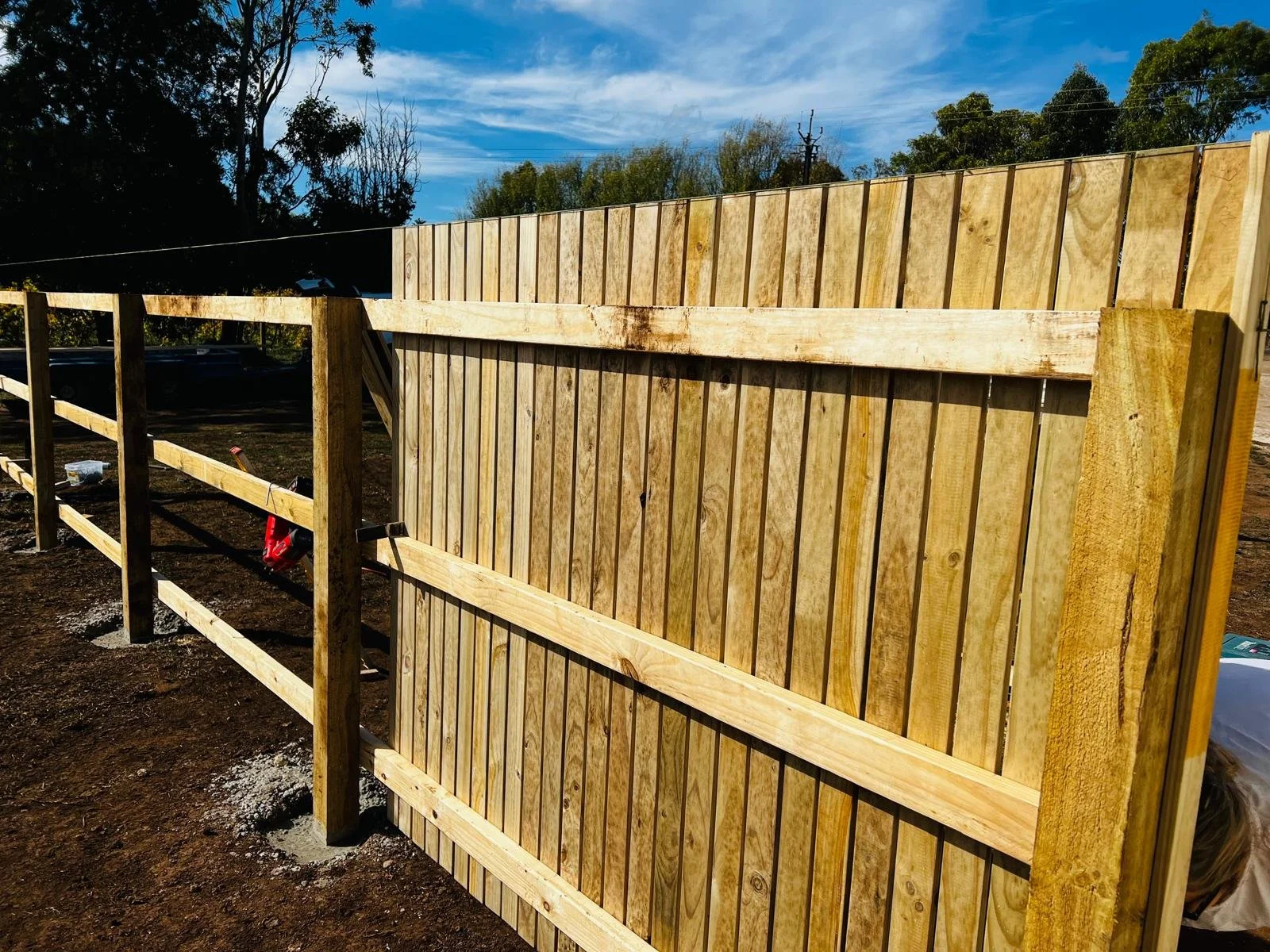 Wooden fence in construction on a dirt ground with trees and blue sky in the background.