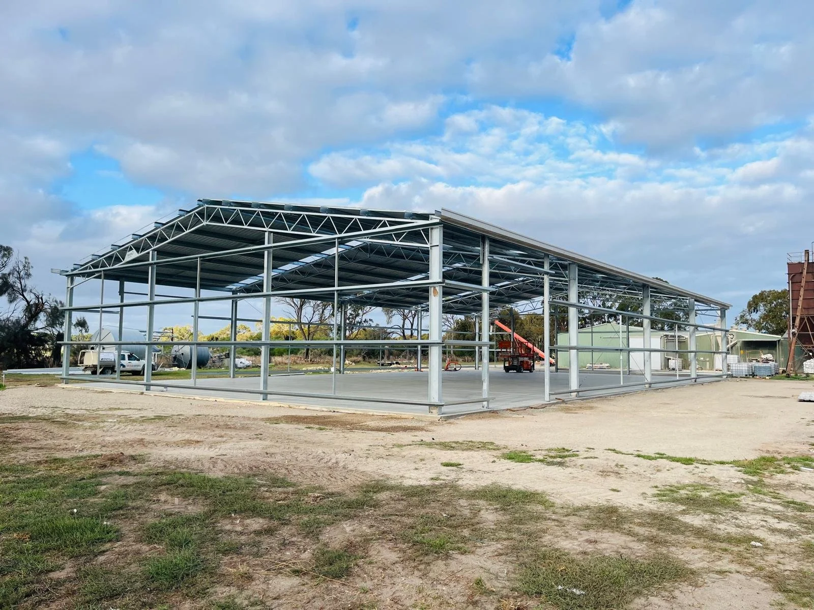The image shows a steel frame structure of a building under construction with a metal roof partially installed. There are construction vehicles and equipment nearby, and the area is open with a partly cloudy sky overhead.