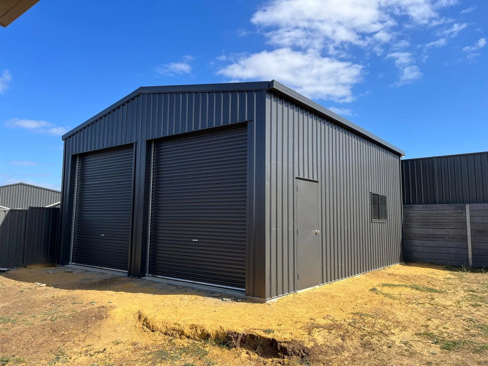 A gray metal storage shed with a large roll-up door, a smaller side door, and a window, situated on a patch of dirt with a blue sky and clouds overhead.