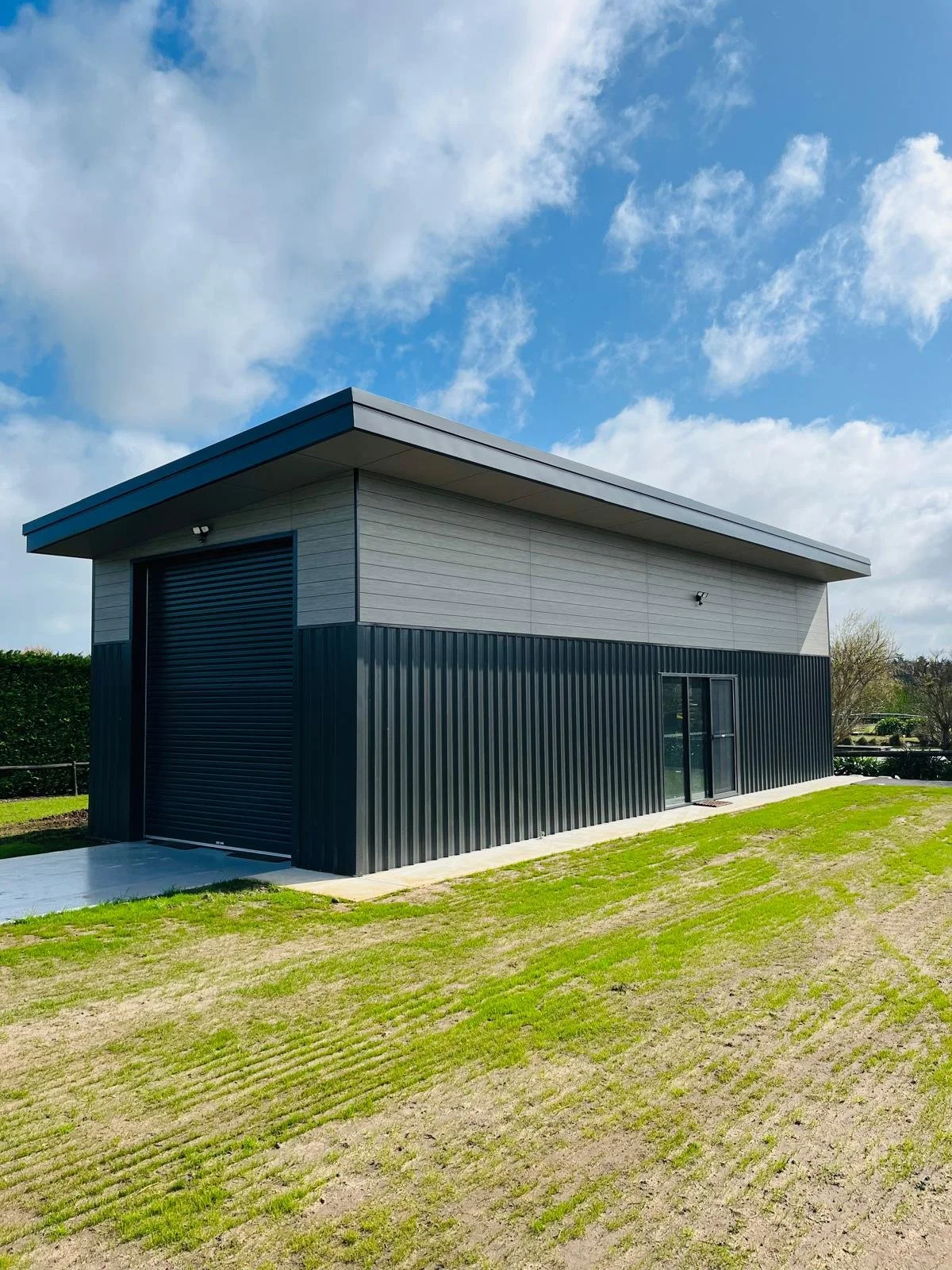 A modern two-story building with a black garage door and a sliding glass door on a grassy yard, under a partly cloudy sky.