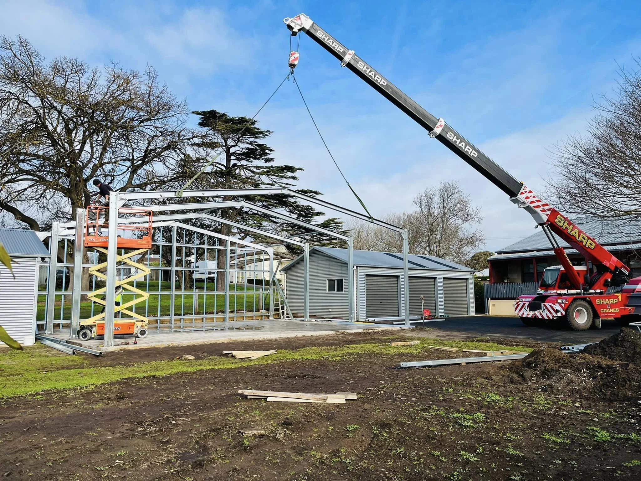 Construction workers are assembling a building frame using a crane and scaffolding in a residential area with trees and houses.