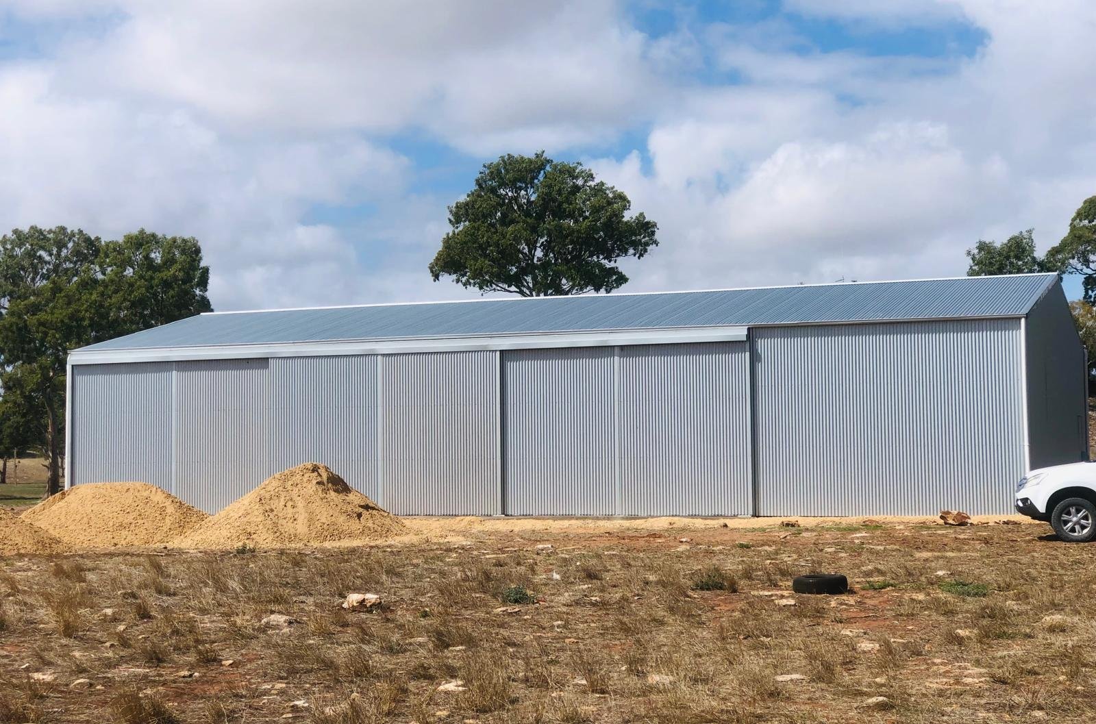 A large, silver metal shed with a sloped roof, situated outdoors with a few piles of dirt and a white vehicle nearby, under a partly cloudy sky with trees in the background.