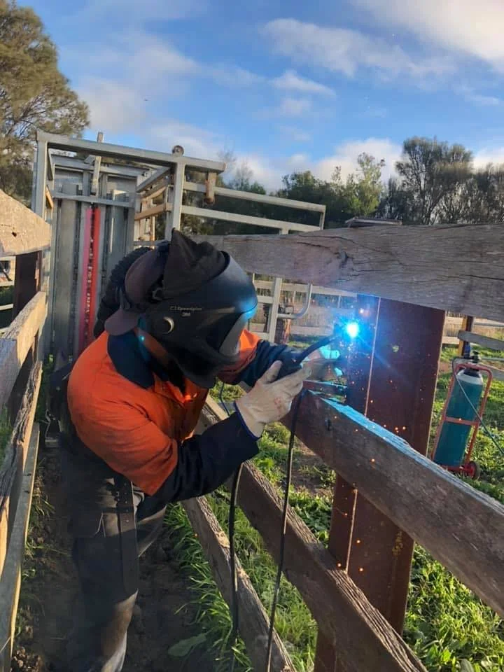 A person welding a wooden fence outdoors during daytime, wearing protective gear including a helmet, gloves, and an orange jacket, with sparks flying from the welding process.