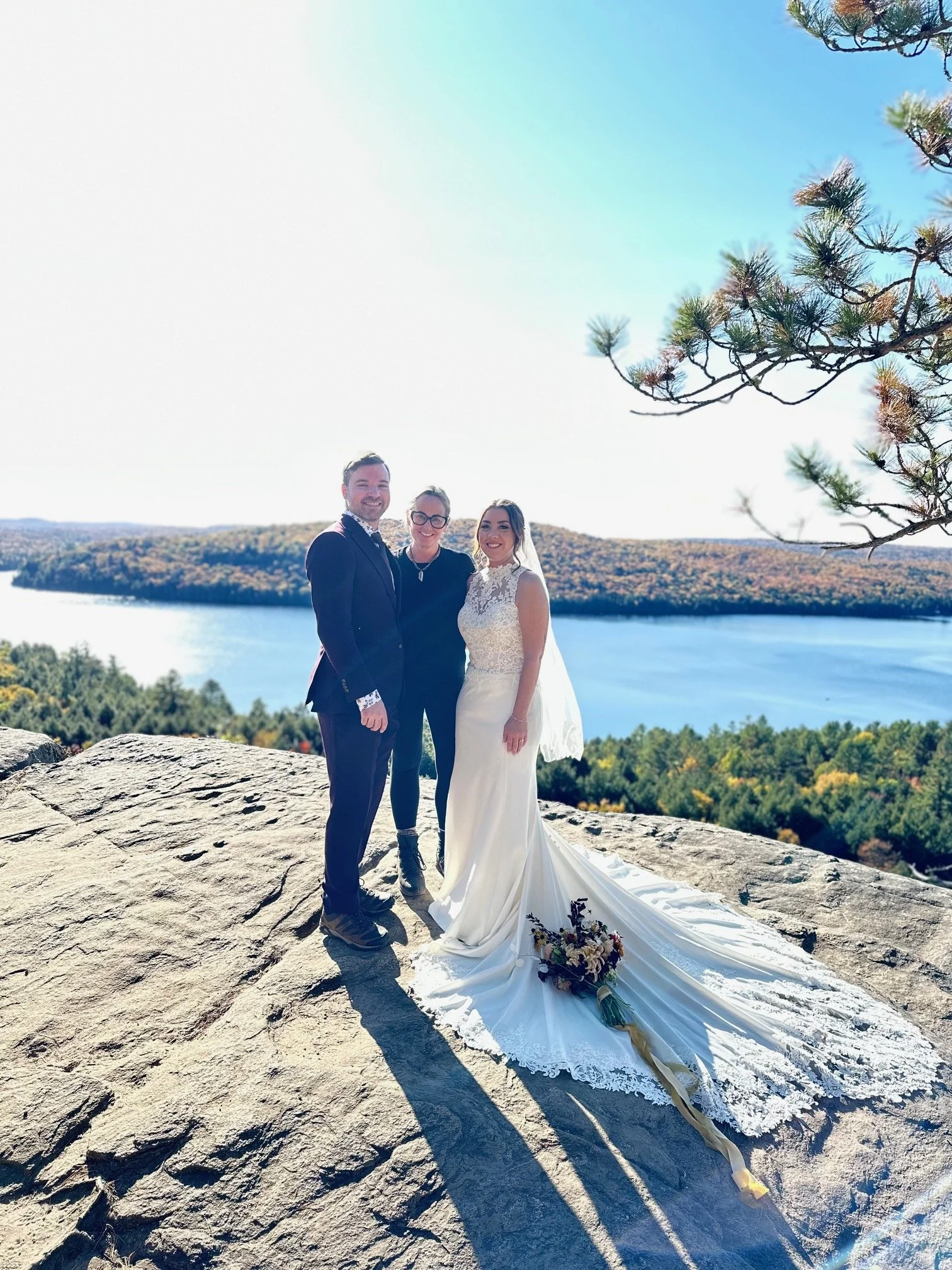 A bride and groom with a woman, probably the officiant, standing together on a rocky outcrop overlooking a lake and forests, with clear blue sky overhead. The bride is in a white wedding gown with a train and bouquet, the groom is in a dark suit, and