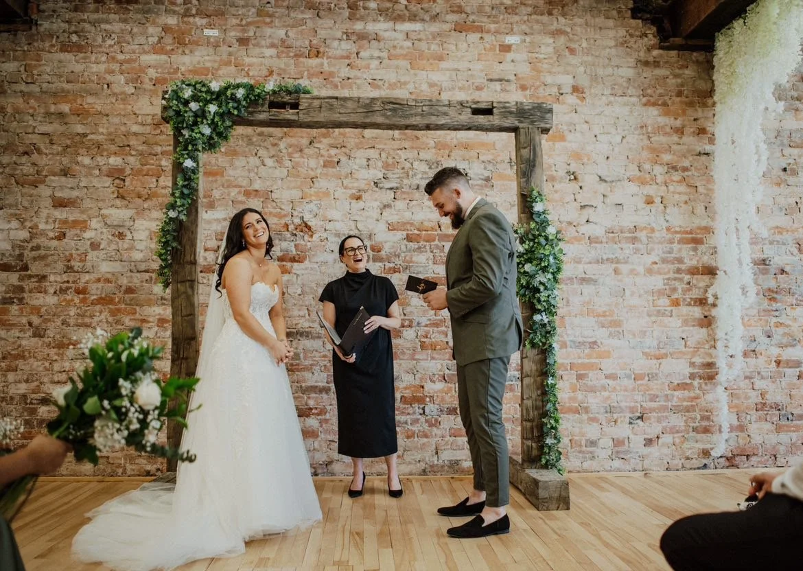 A wedding ceremony with a bride and groom exchanging vows under a rustic wooden arch decorated with green foliage and white flowers in front of a brick wall. The bride is in a white wedding dress, and the groom is in a grey suit. An officiant in a bl