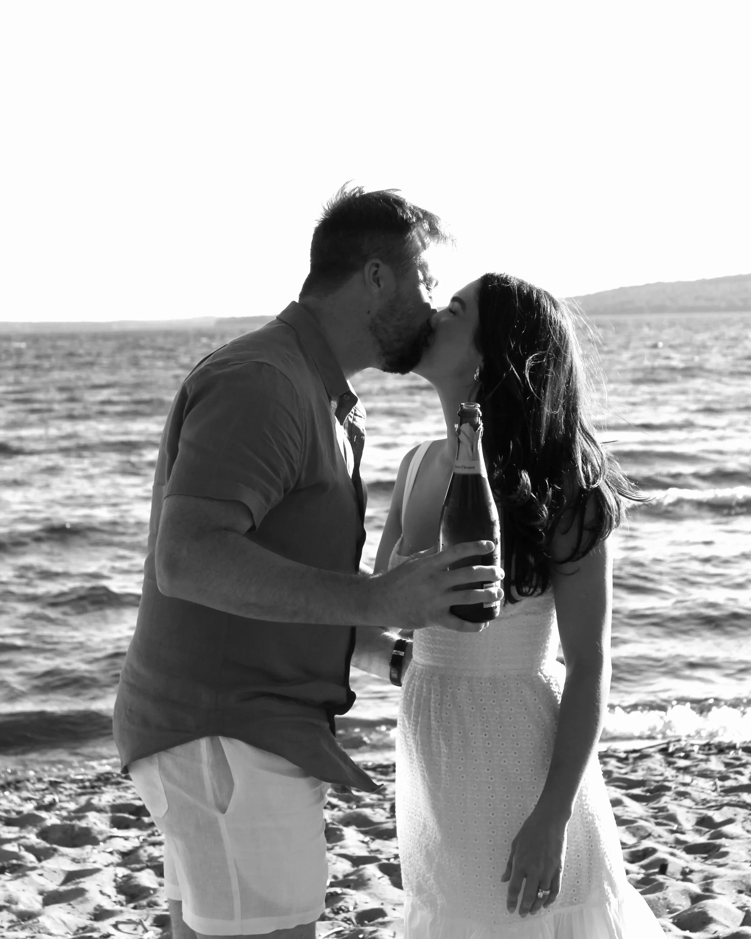 A couple kissing on the beach, with the man holding a bottle of beer, in black and white.