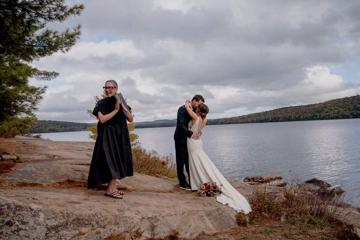 A wedding ceremony taking place outdoors near a lake with the bride and groom kissing, and the officiant standing nearby smiling.