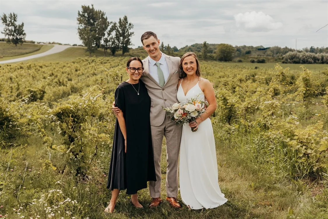 A bride, groom, and two women standing in a vineyard, smiling, with the bride holding a bouquet of flowers.