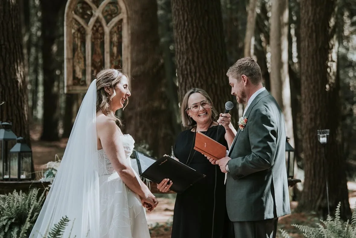 A wedding ceremony in a forest with a bride and groom smiling at each other, an officiant holding a microphone, and holding a book, during their vows.