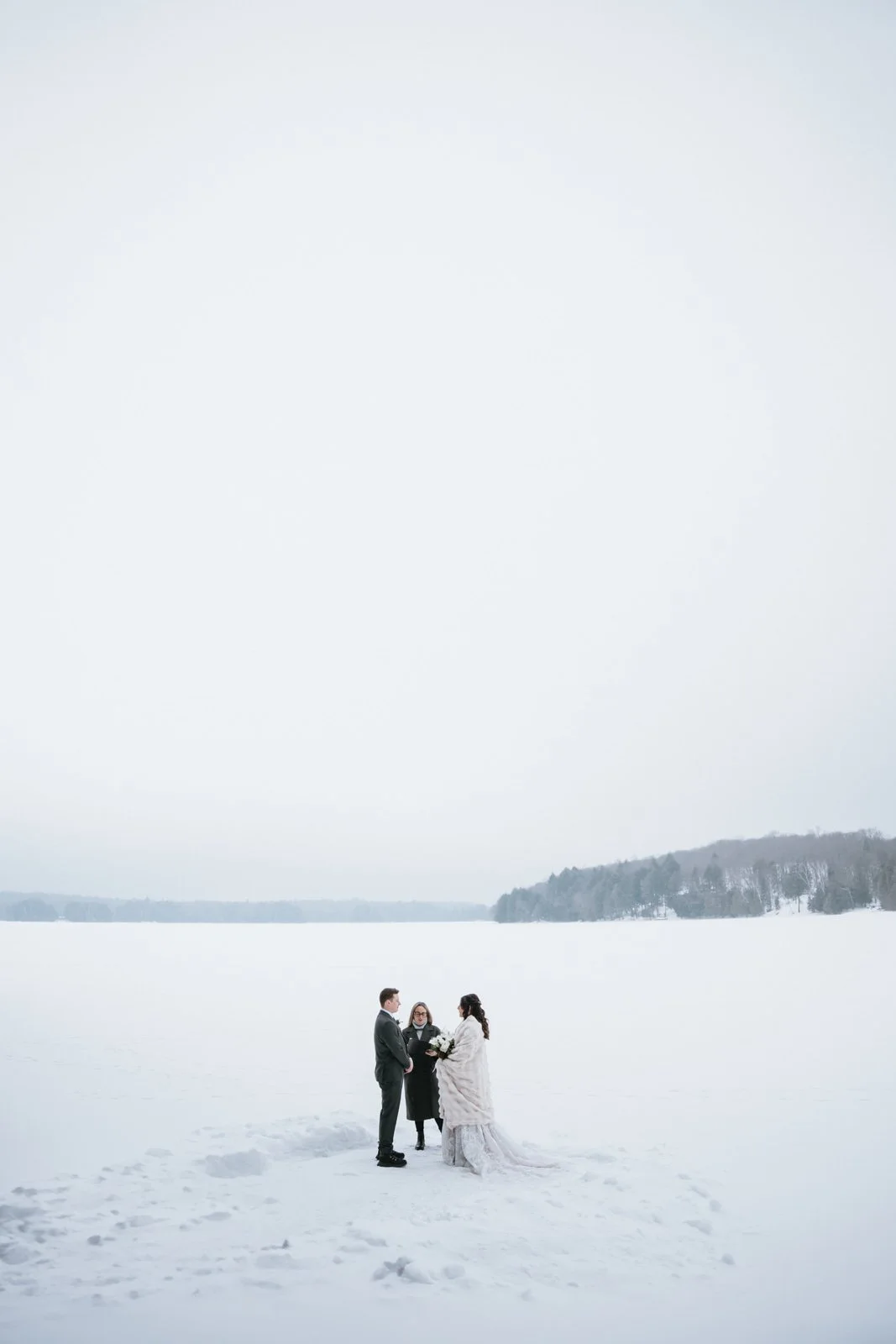 A bride and groom standing in the snow by a frozen lake, facing each other with an officiant between them, during a winter wedding.
