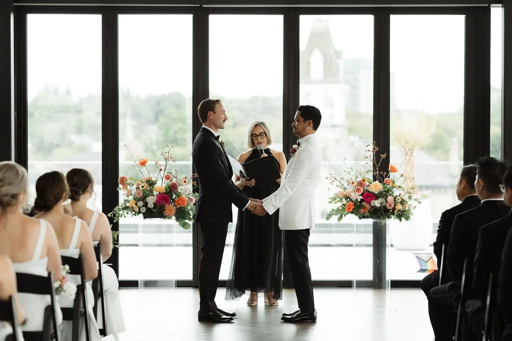 A couple getting married, holding hands and facing each other, with an officiant and a large window showing an outdoor cityscape background. Guests are seated on either side, with floral arrangements on stands.