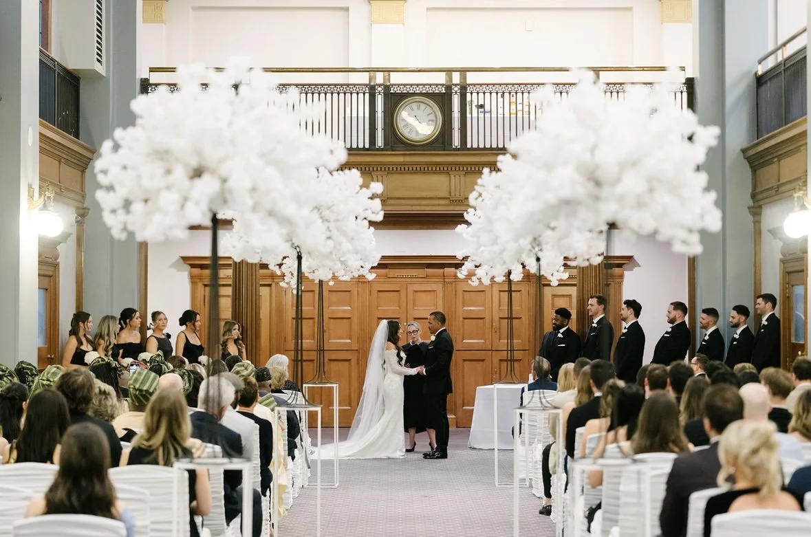 Bride and groom exchange vows in a wedding ceremony at an indoor venue with large white floral decorations and an audience of guests.