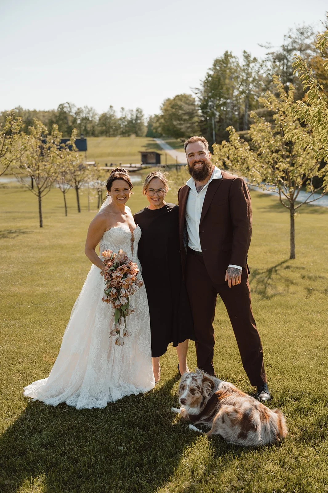 A bride in a white wedding dress holding a bouquet, standing next to a woman in black, and a man in a dark suit, with a dog laying on the grass in front, in a sunny outdoor setting with trees and a small building in the background.