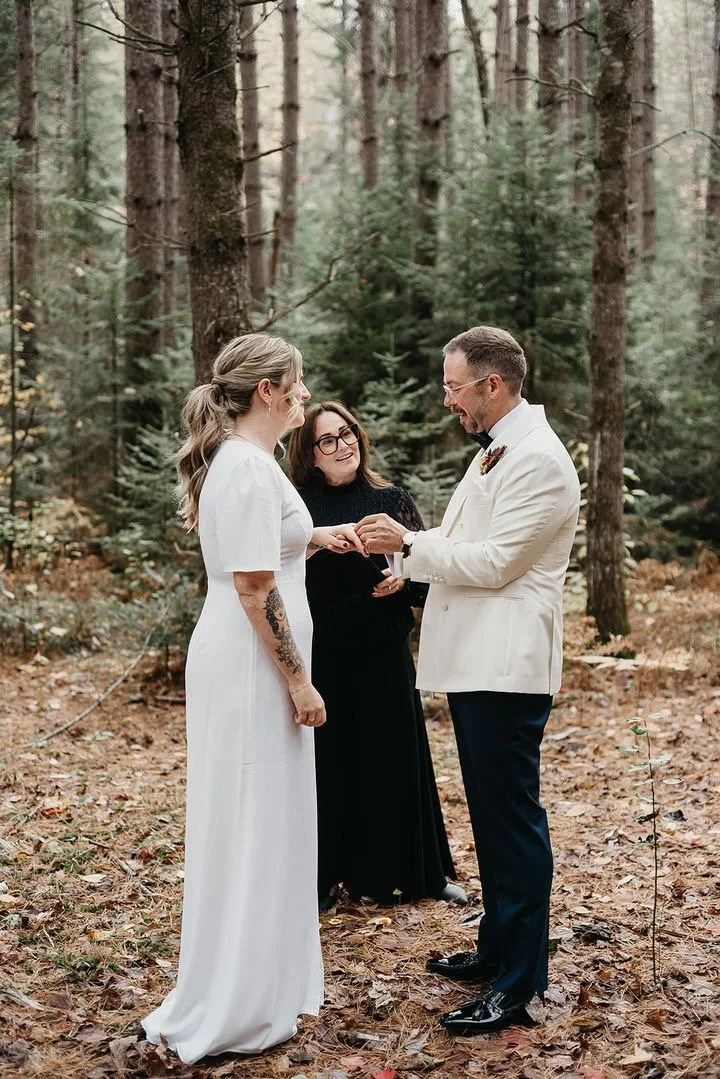 A couple getting married in a forest, exchanging vows, with officiant standing behind them, surrounded by tall trees and fallen leaves.