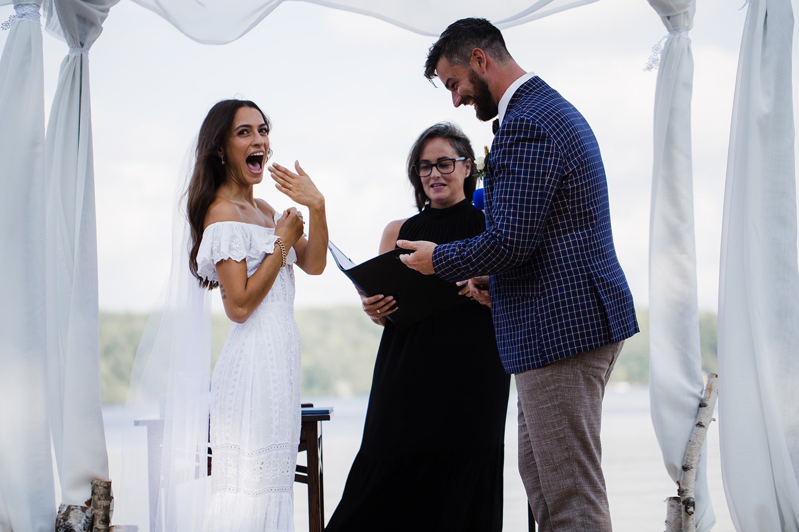 A bride with long dark hair, wearing a white off-the-shoulder wedding dress, is smiling and reacting with surprise at her wedding ceremony. The officiant, a woman in a black dress, is holding a booklet, while the groom, dressed in a checkered suit, i
