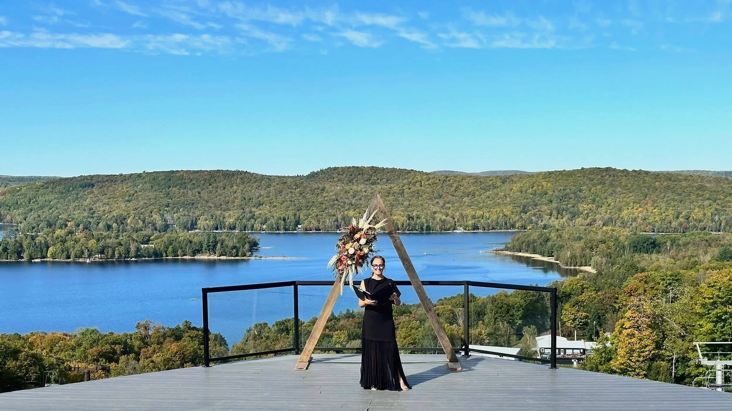 A woman in a black dress standing under a decorated wooden arch on a rooftop with a lake and forested hills in the background.