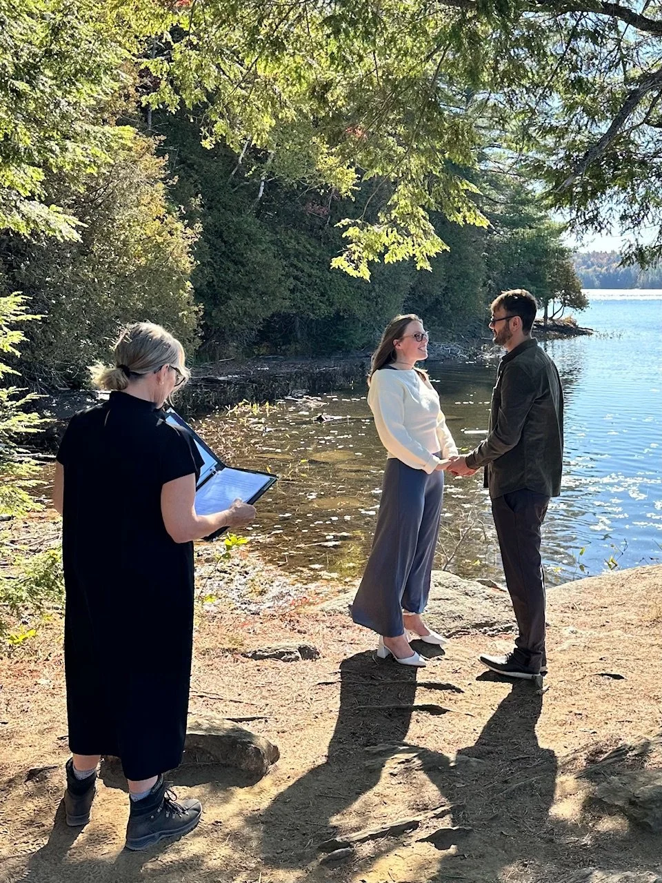 A couple holding hands and exchanging vows on a tree-lined lakeshore during a wedding ceremony, with a woman officiant reading from a book nearby.