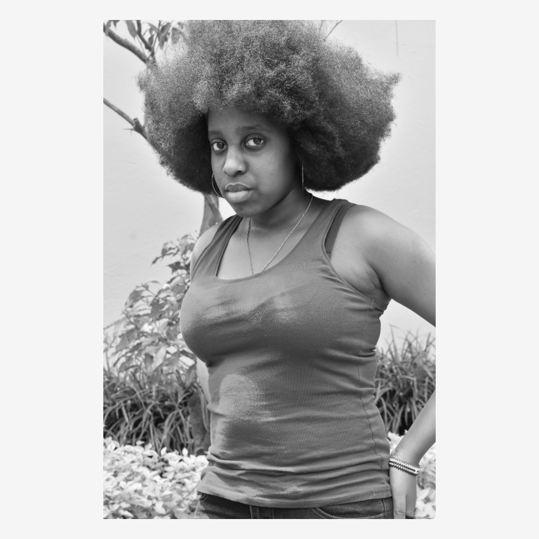 Black and white photo of a woman with large, curly afro hair, wearing a sleeveless top, earrings, wrists accessories, and a necklace, standing outdoors with plants in the background.