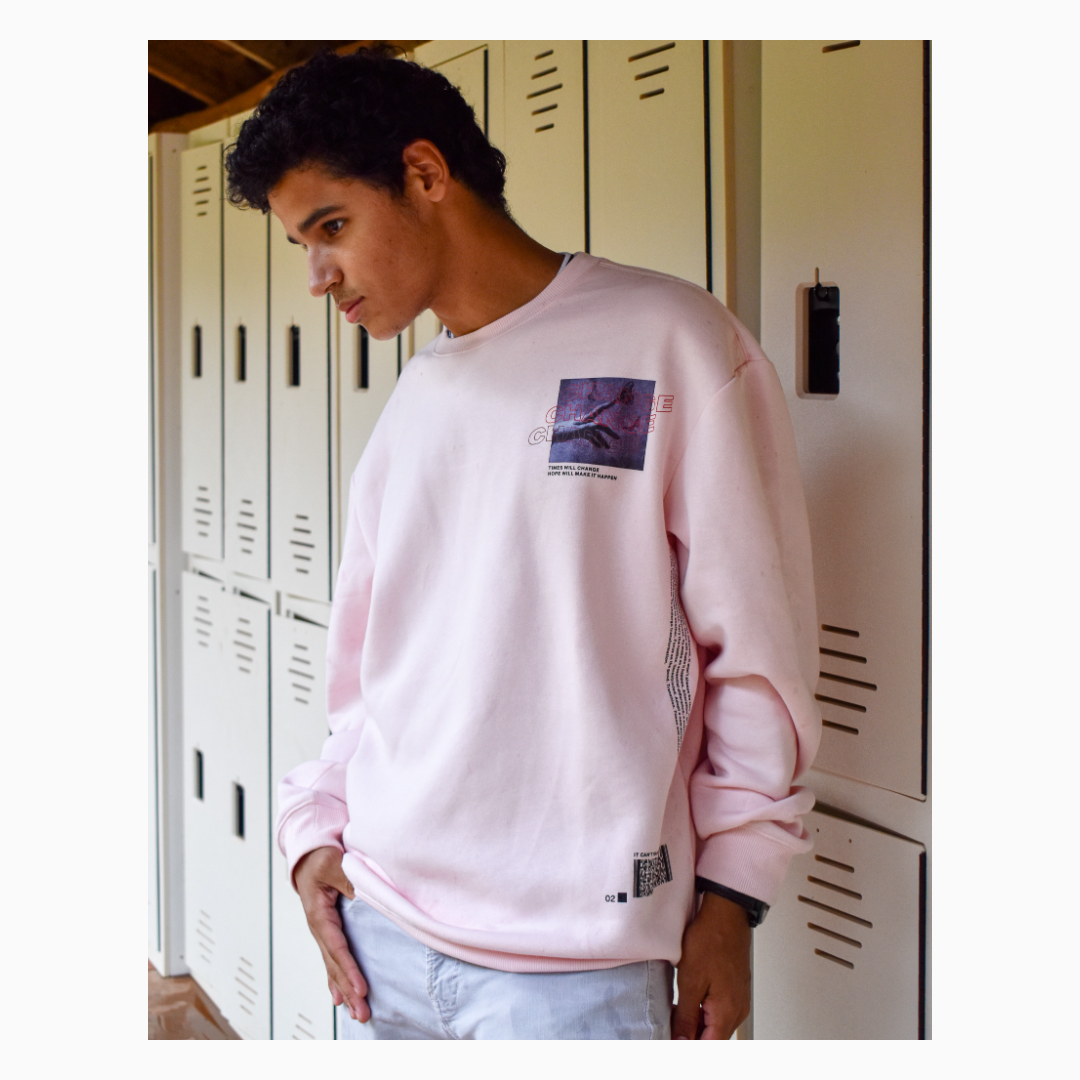 A young man with dark curly hair leaning against lockers, wearing a light pink sweatshirt with graphic patches and light-colored jeans.