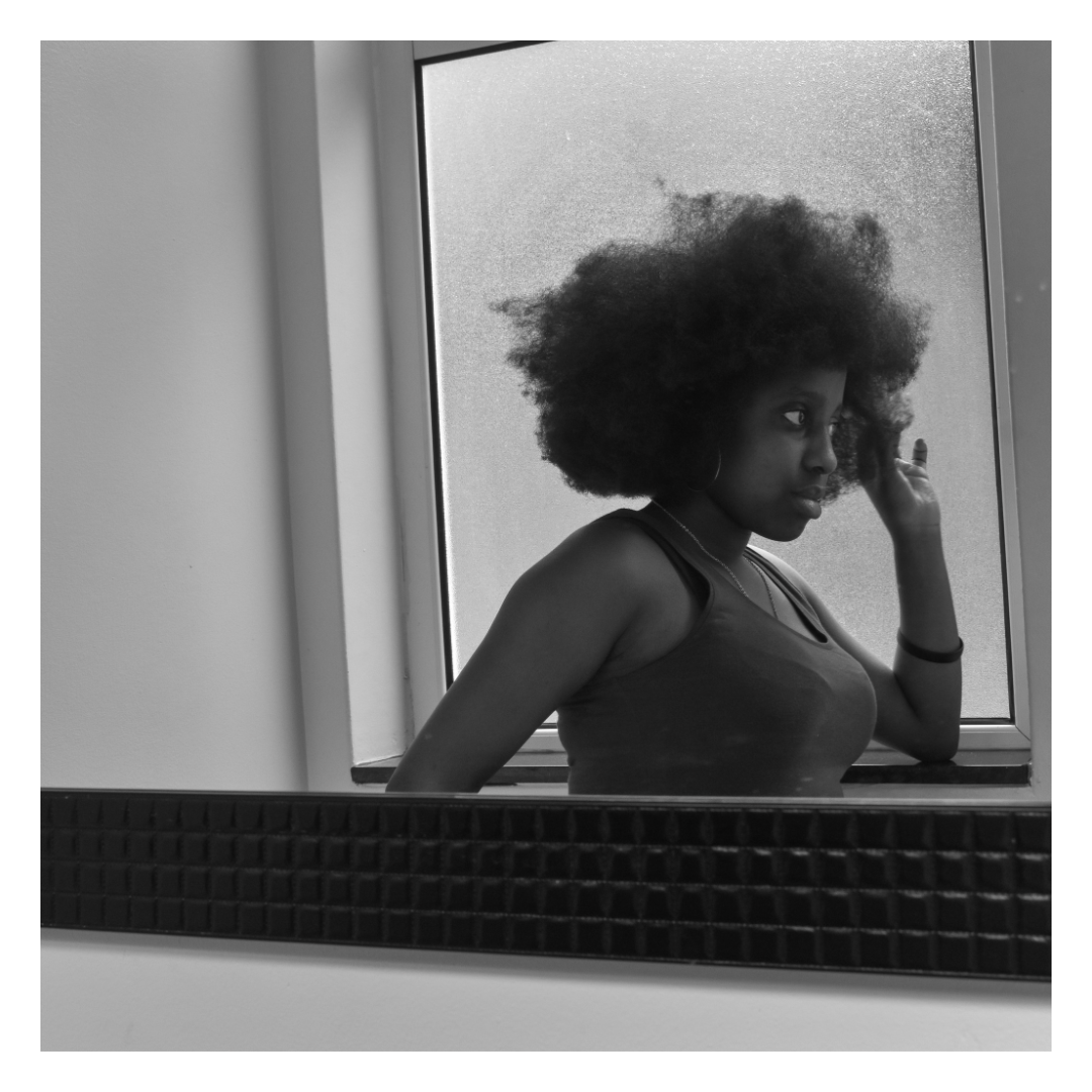 A Black and white photo of a woman with large natural curly hair looking into a mirror. She is wearing a sleeveless top and earrings, and is touching her hair with her right hand. The photo is taken through a window or mirror with a textured black fr