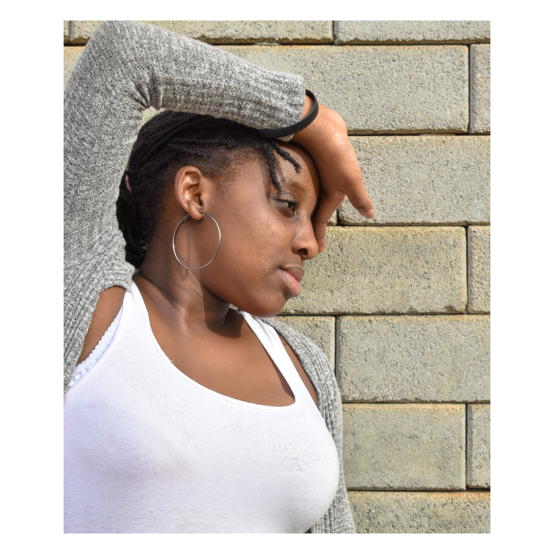A woman with dark skin, wearing a white top and large hoop earrings, leans against a textured beige brick wall with her eyes closed and one arm resting on her forehead.