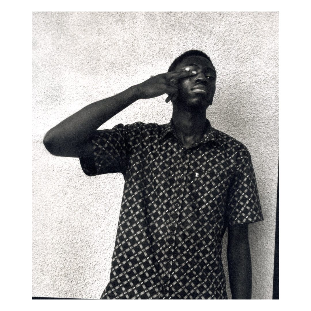 A young man standing against a textured wall, saluting with his right hand, wearing a patterned short-sleeve shirt.