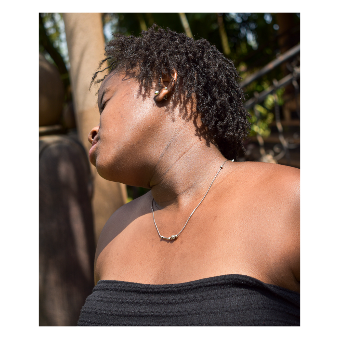 Close-up of a woman with short curly hair, wearing a silver necklace and small earrings, outdoors with greenery in the background.