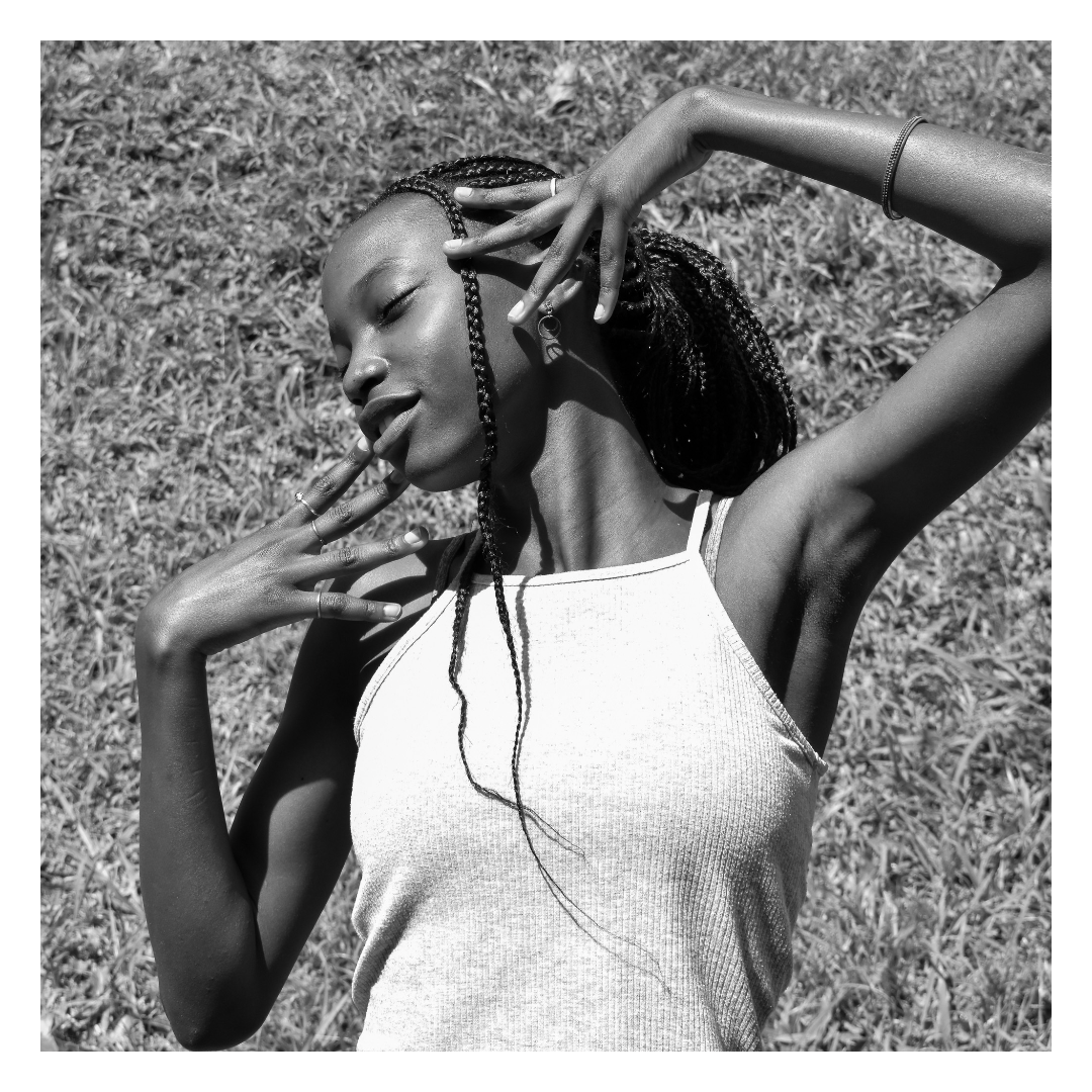 A black and white photo of a woman with braided hair lying on grass with her eyes closed, posing with her hands near her face and neck, wearing a sleeveless top and jewelry.