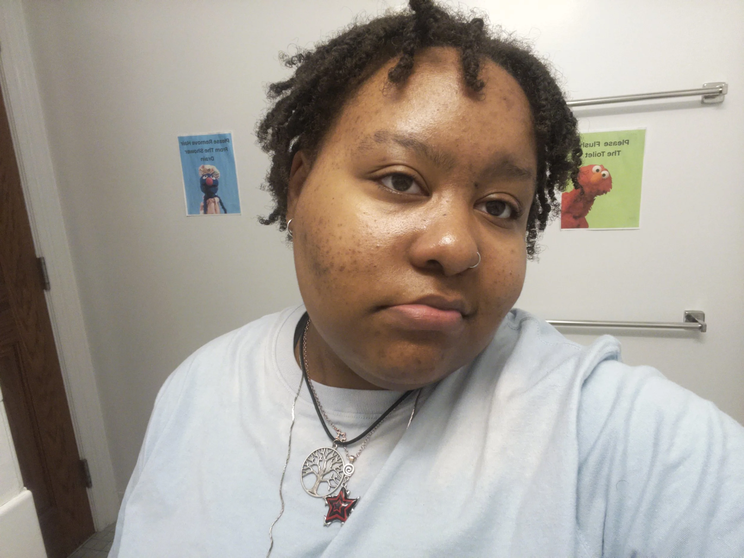 A person with short, curly hair and a nose piercing, wearing a light-colored shirt and layered necklaces, taking a selfie in a room with white walls and humorous posters in the background.