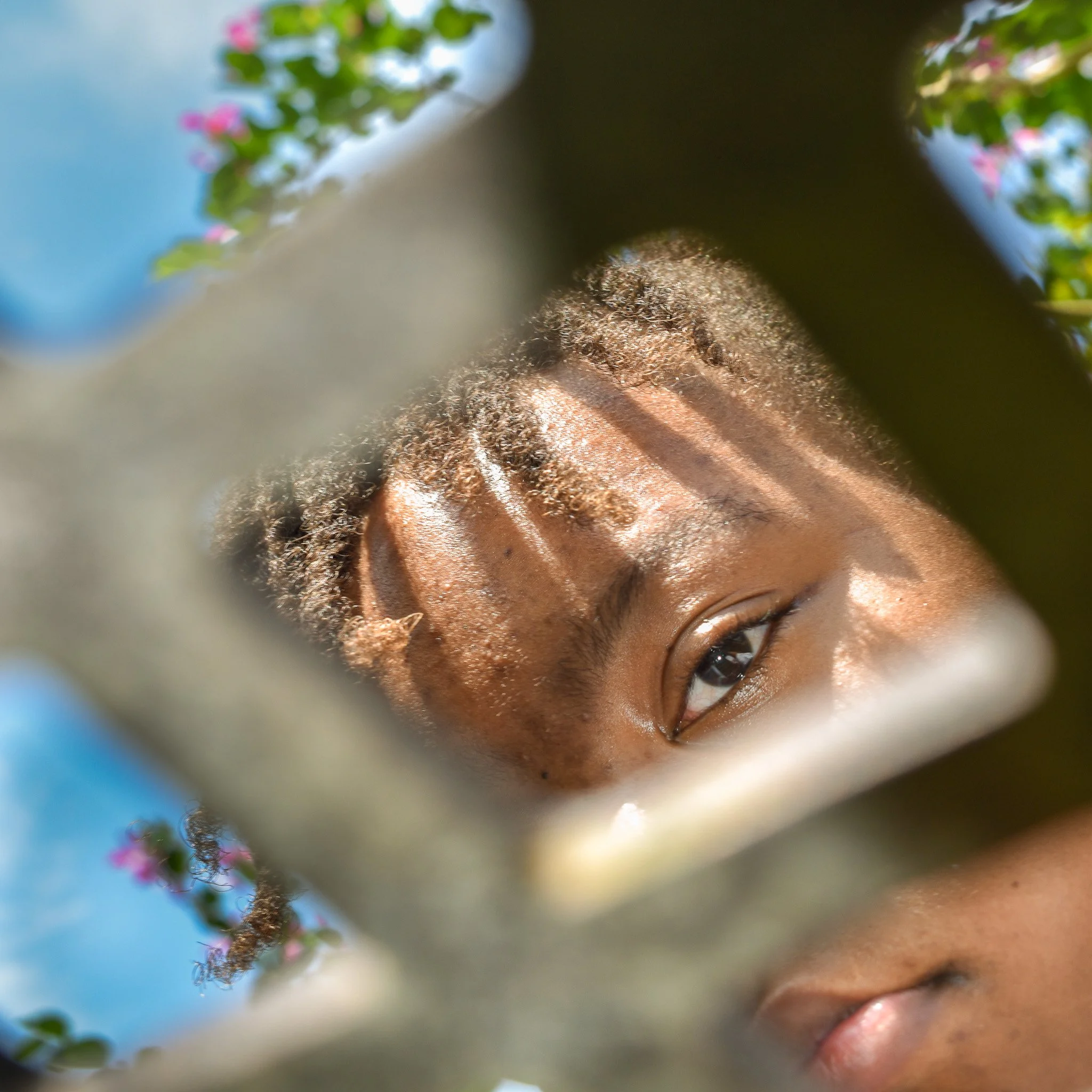 Close-up of a young person's face with one eye visible, framed by leaves and pink flowers, with sunlight casting shadows.