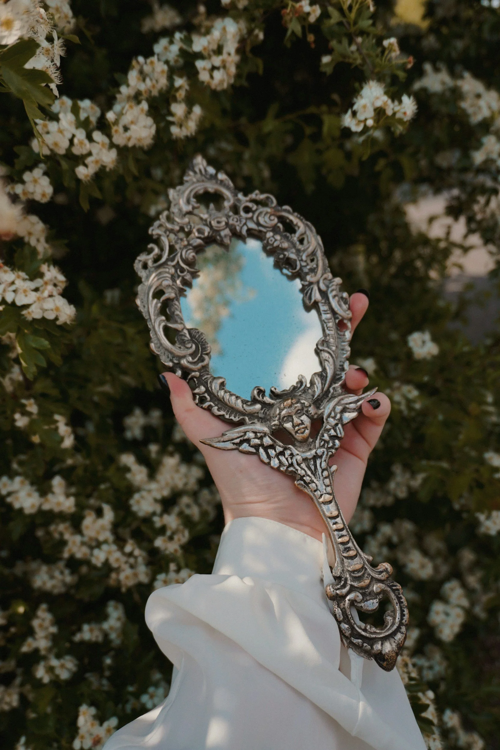 hand holding an ornate mirror with white flowers in the background representing existential crisis of an attractive woman who is aging