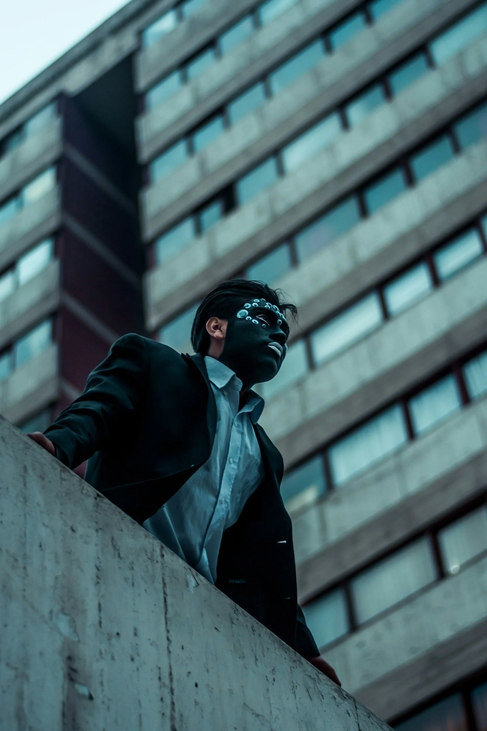 Young adult male dressed business casual and wearing a mask stands on a rooftop in an urban setting surrounded by high rise office buildings representing imposter syndrome