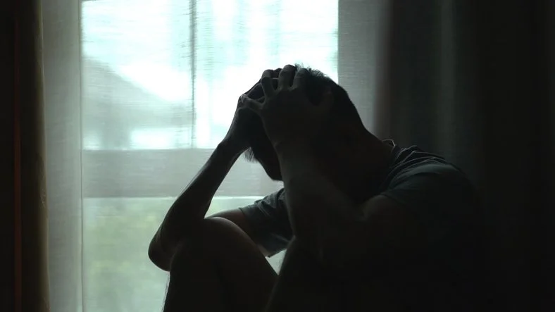 Man with hands to his head in a darkened room by a window, signifying burnout