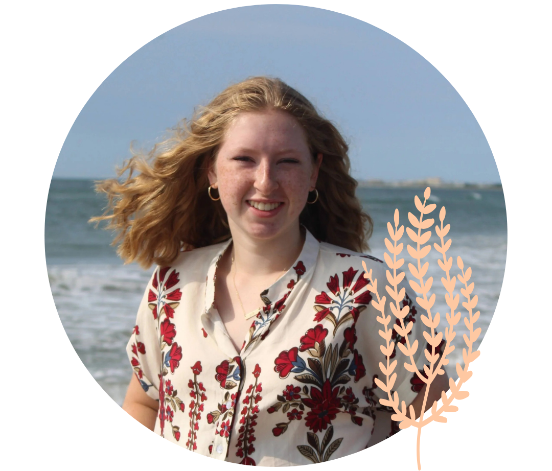 Young woman with red hair, wearing a floral blouse, standing on a beach with waves in the background.