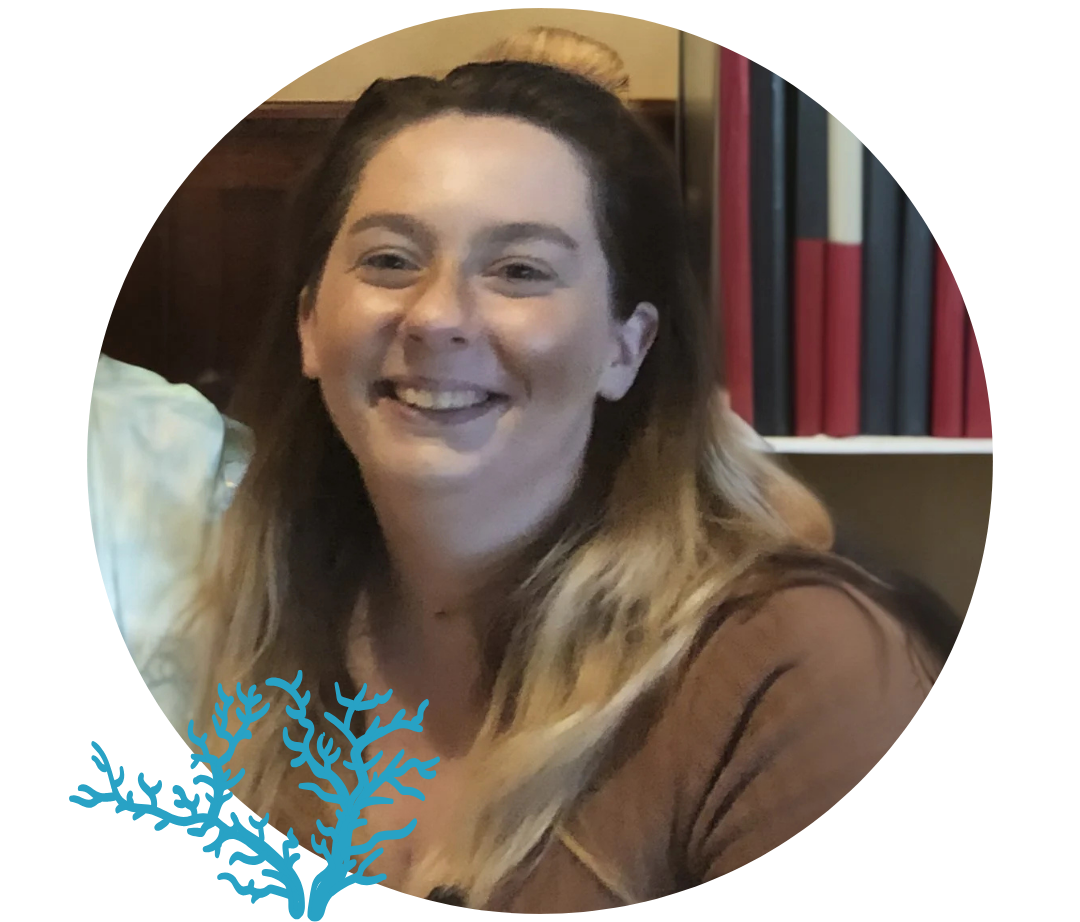 A smiling woman with long brown hair and light skin, sitting indoors near a bookshelf with colored books.