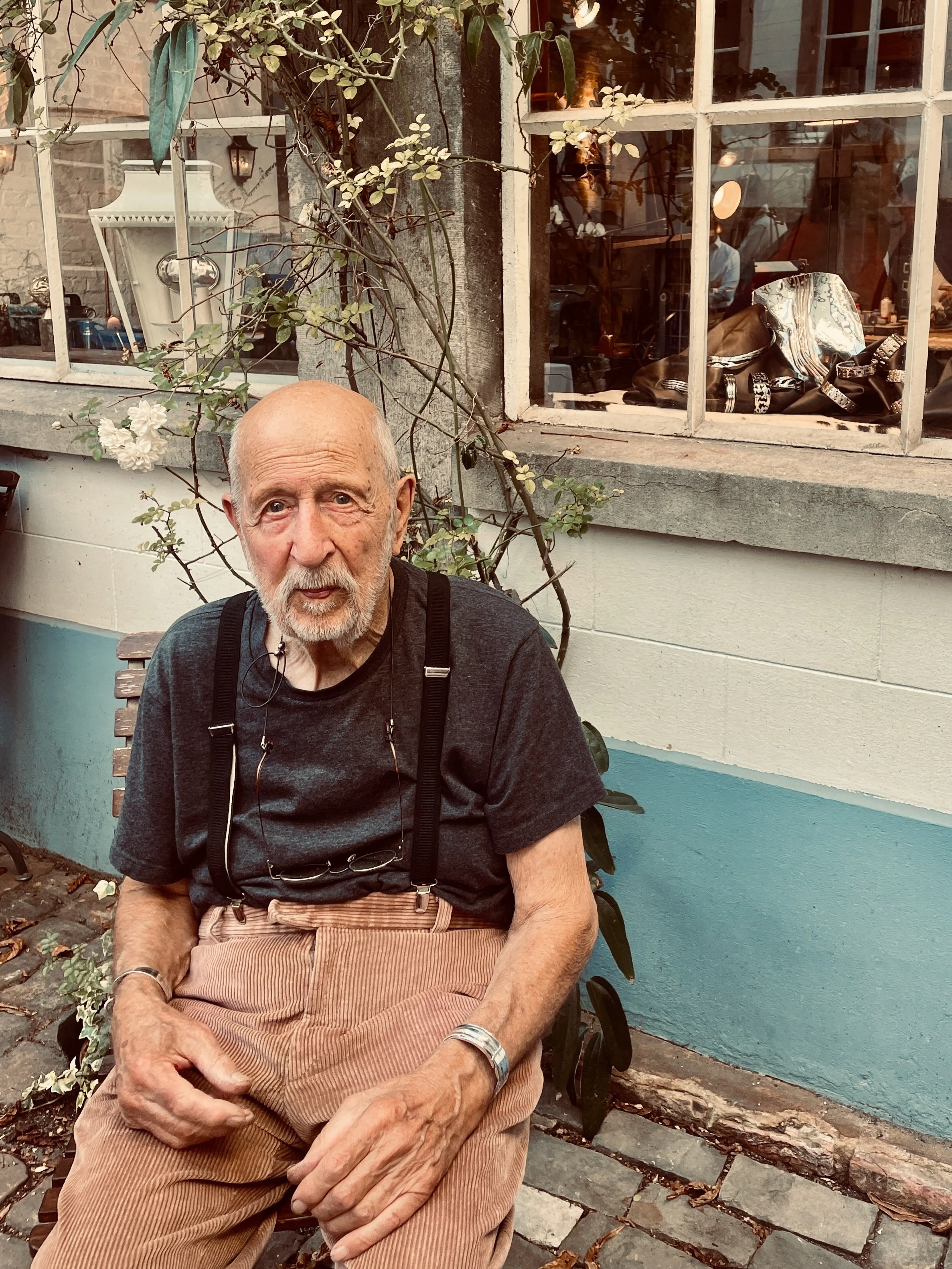Jacques Moniquet assis sur un banc en extérieur, portant un t-shirt noir, un pantalon beige à rayures, des suspenders, et une montre. En arrière-plan, il y a une maison avec une fenêtre et des plantes, y compris une branche avec des fleurs blanches.