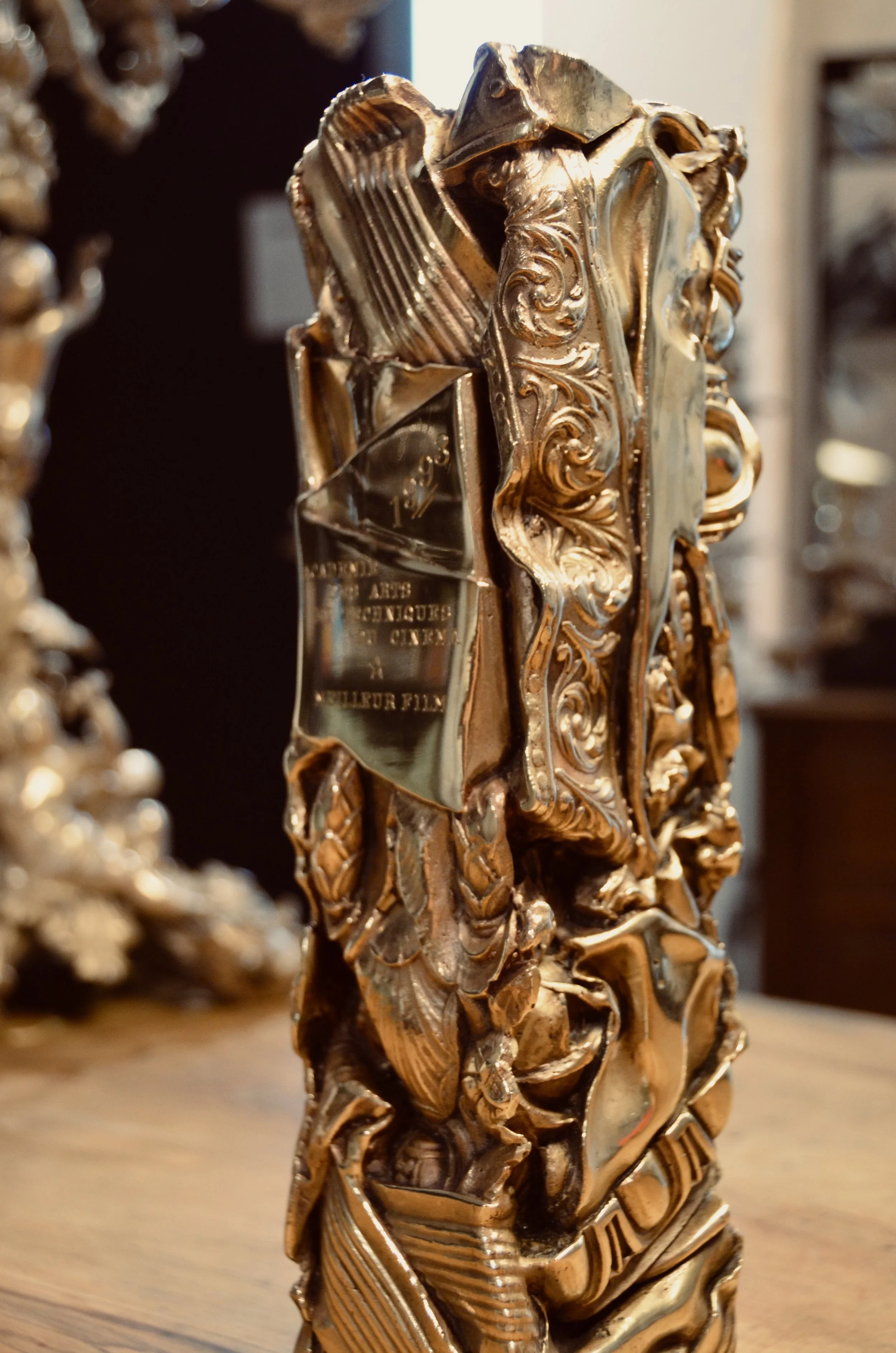 Une sculpture ornementale en bronze patiné avec des détails sculptés complexes, probablement une pièce décorative ou un trophée, placée sur une surface en bois.