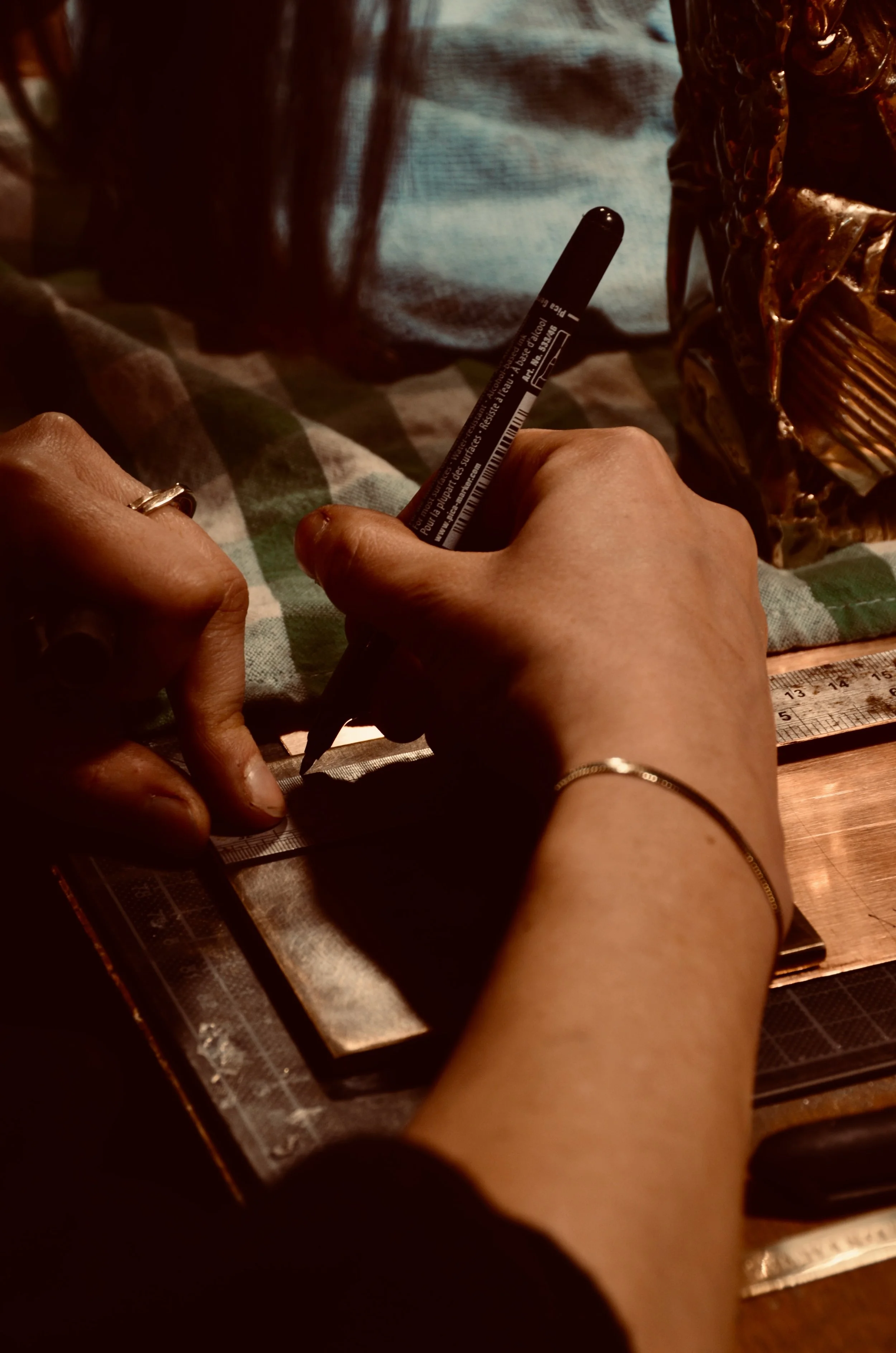 Une personne qui utilise un stylo noir pour faire une marque sur une règle en métal sur une surface en bois. L'image montre aussi une main portant une bague et un bracelet, avec un tissu à carreaux en dessous.