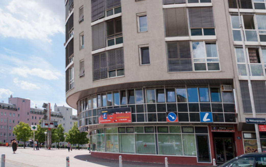 Ein mehrstöckiges Bürogebäude mit einer Ecke in einer Stadt, im Vordergrund ein Platz mit wenigen Menschen, Bäumen und Verkehrszeichen.