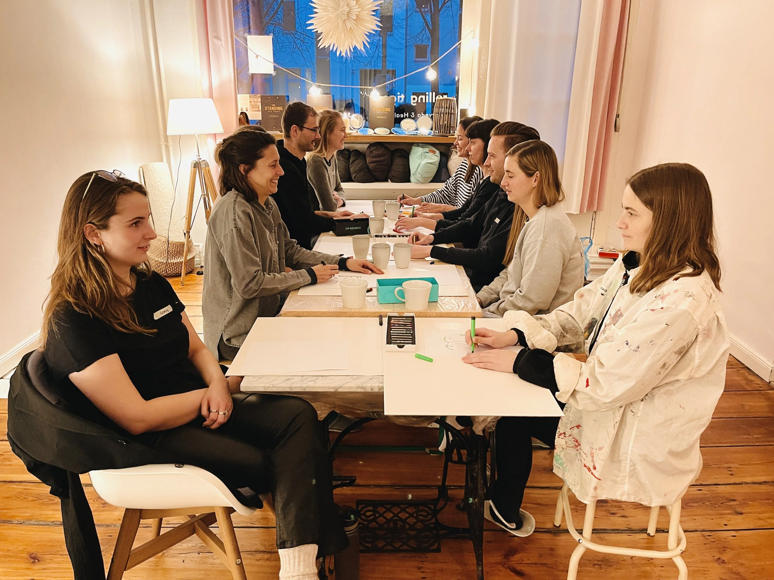 A group of people seated around a long table in a warmly lit room, engaged in a meeting or discussion, with some taking notes and others smiling.