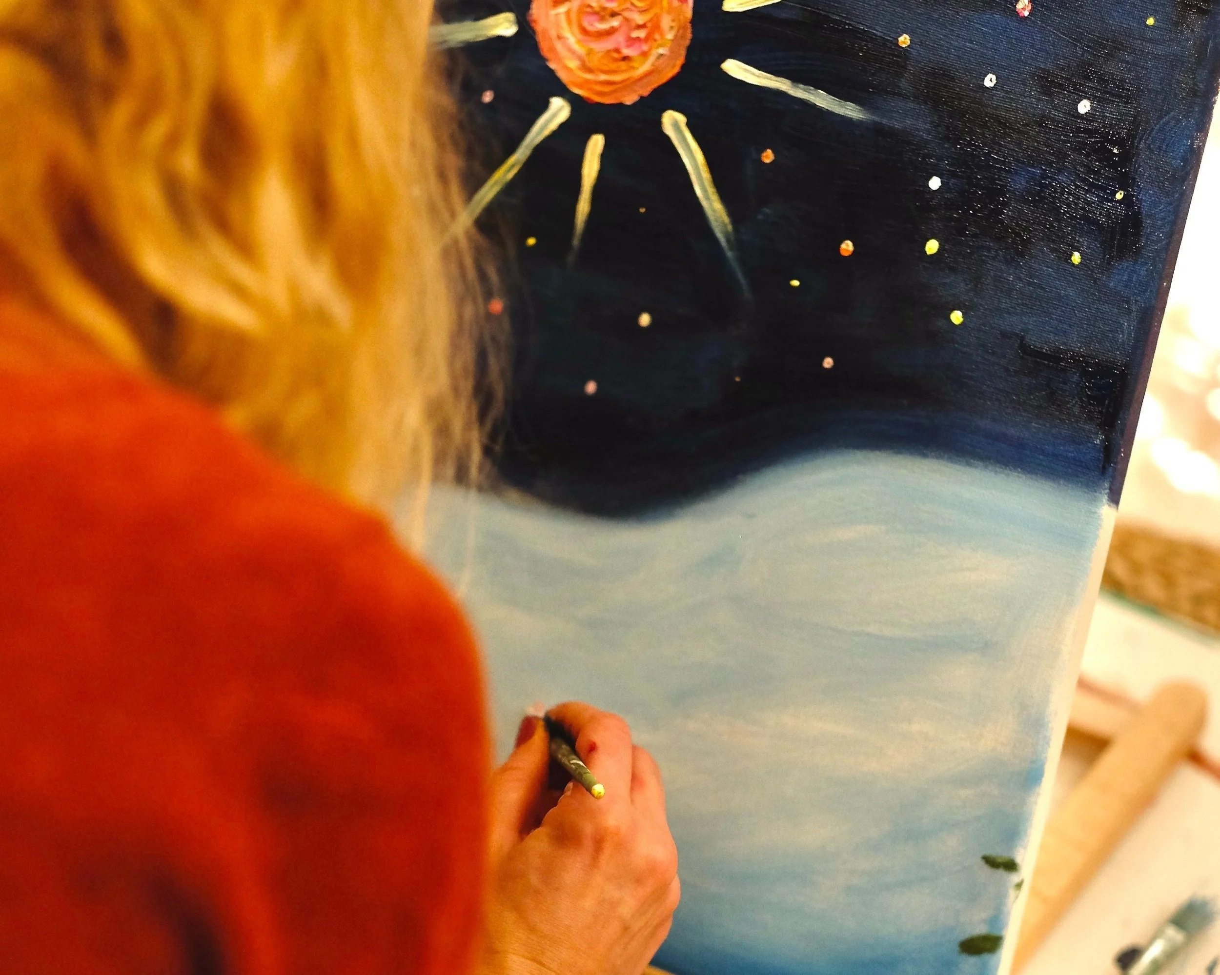 A person with blonde hair and a red top painting a night scene with a large, radiant orange star, streaks of light, and small stars in a dark sky on a canvas.