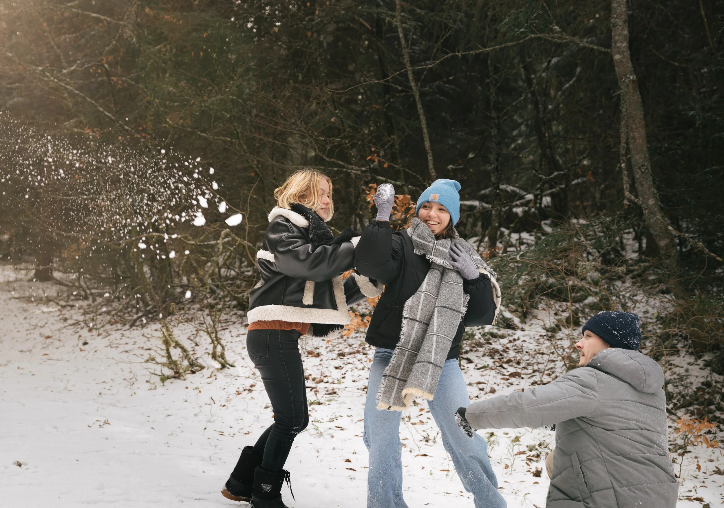 Trois personnes s'amusant dans la neige, habillées avec des vêtements chauds, dans une forêt enneigée.