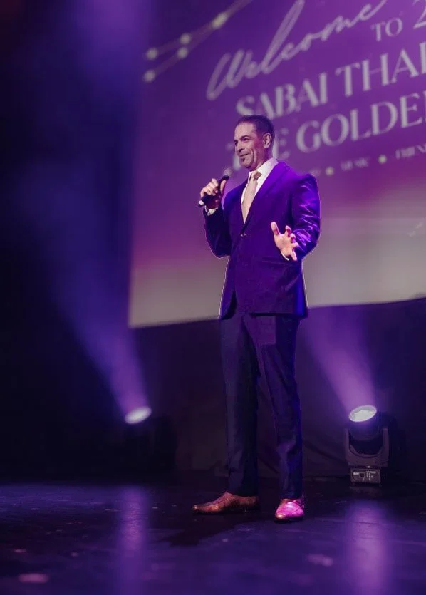 A man in a purple suit and brown shoes standing on stage, holding a microphone, with purple stage lighting and a screen in the background displaying text.